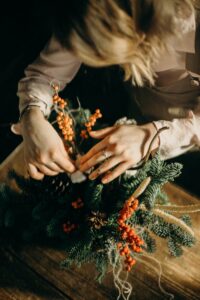 Hands arranging a festive Christmas wreath with pine and red berries on a wooden table.